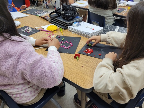 Two students are seated at a table. They have black construction paper in front of them and are placing flower petals on this paper.