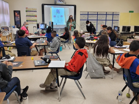 Classroom full of elementary students. There is a teacher at the front of the classroom by a projector screen. The screen has an image of a flower diagram on it.