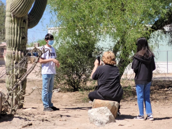 One middle school student is standing in the distance. One student sits on a rock holding their phone to film the first student. There is one additional student standing next to the one filming.