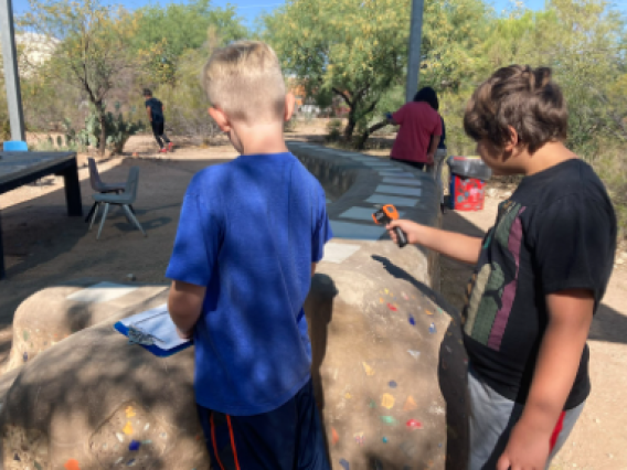 Picture shows two students recording temperature data from the ground outdoors. One male student on the left is wearing blue and one male student on the right is wearing read and holding a thermometer.