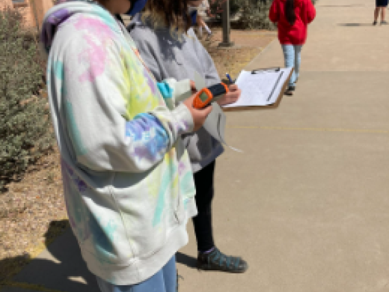 Image shows two students standing on a walkway. The student closest to the camera is wearing a tie-dye hoodie and is holding a thermometer. The student farthest from the camera is wearing a grey sweatshirt and is writing on a piece of paper that is on a clipboard.