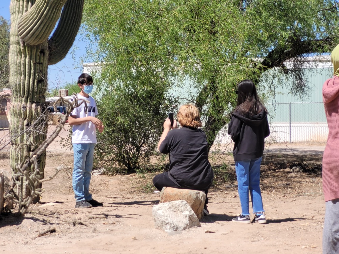 One middle school student is standing in the distance. One student sits on a rock holding their phone to film the first student. There is one additional student standing next to the one filming.