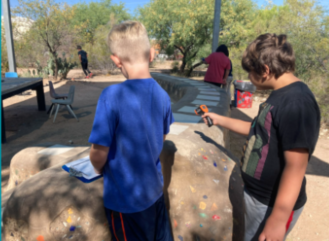 Picture shows two students recording temperature data from the ground outdoors. One male student on the left is wearing blue and one male student on the right is wearing read and holding a thermometer.