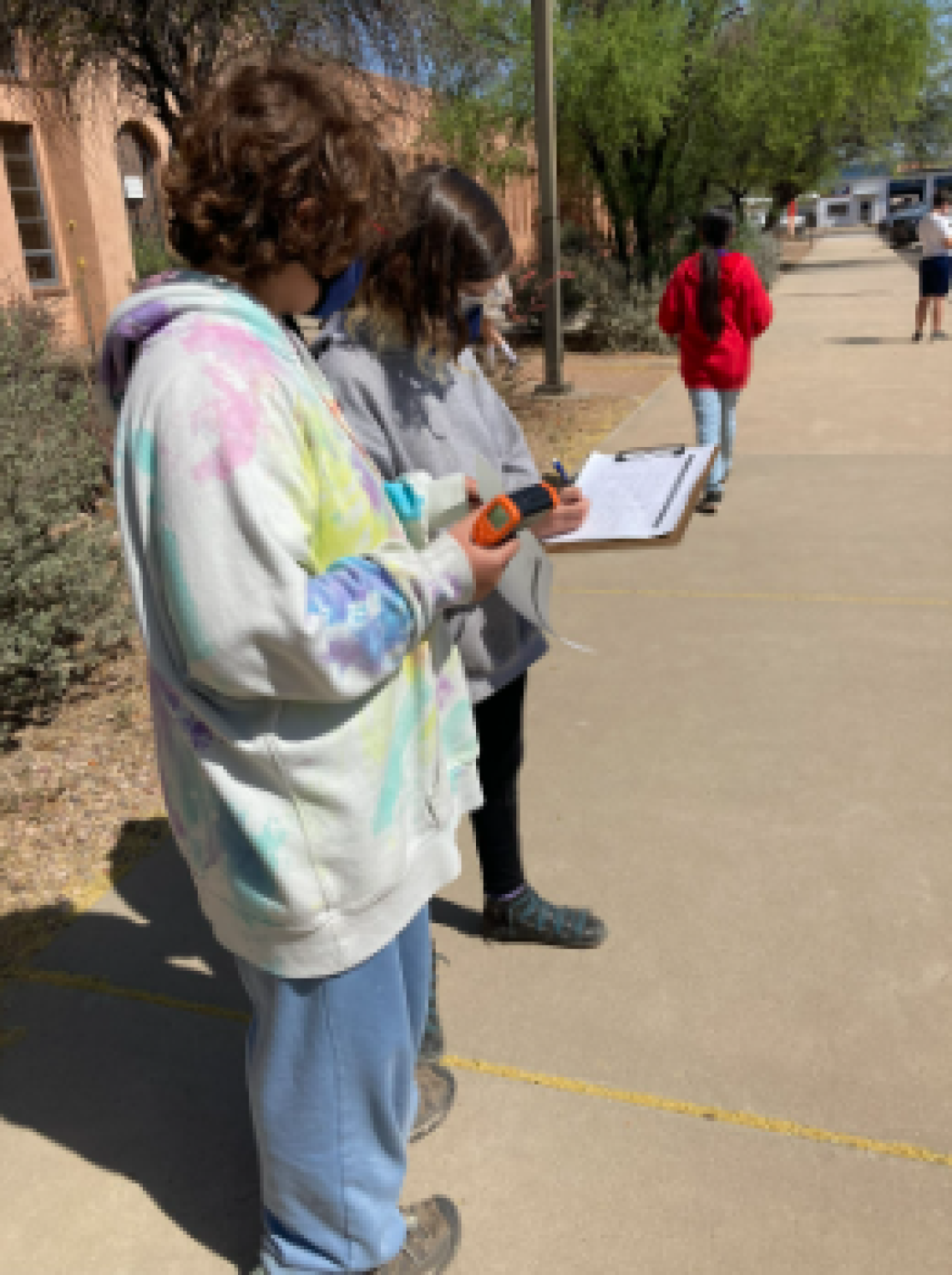 Image shows two students standing on a walkway. The student closest to the camera is wearing a tie-dye hoodie and is holding a thermometer. The student farthest from the camera is wearing a grey sweatshirt and is writing on a piece of paper that is on a clipboard.