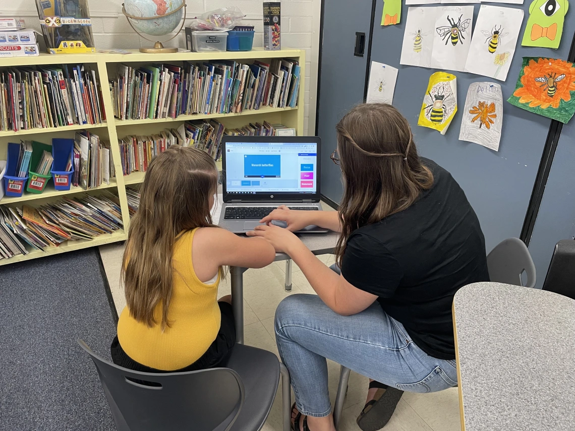 University of Arizona student intern and third grade student are seated next to each other at a desk. The intern is typing on an open laptop in front of them while the student is watching.