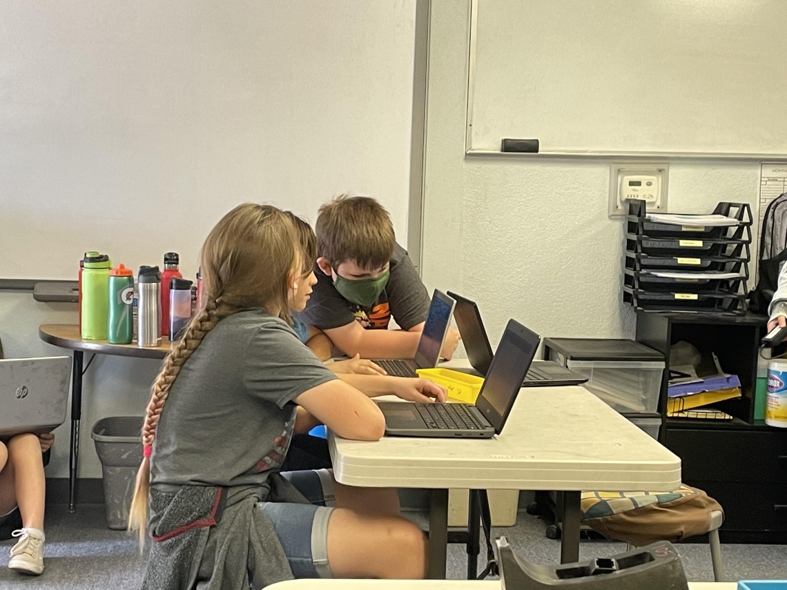 Two fourth grade students are seated at a table with laptops in front of them. One student is standing in on the side of them looking at the laptop screens.